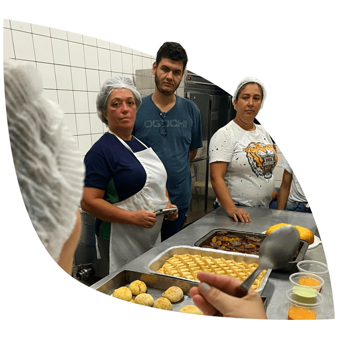 3 alunos (2 mulheres e 1 homem) do Curso Técnico prestando atenção na explicação da professora. Eles estão atrás de uma mesa que contem a produção dos alimentos feitos em sala de aula.